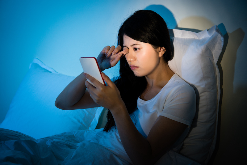 Woman rubbing her eyes feel painful with using smartphone sitting on bed sleeping at night.